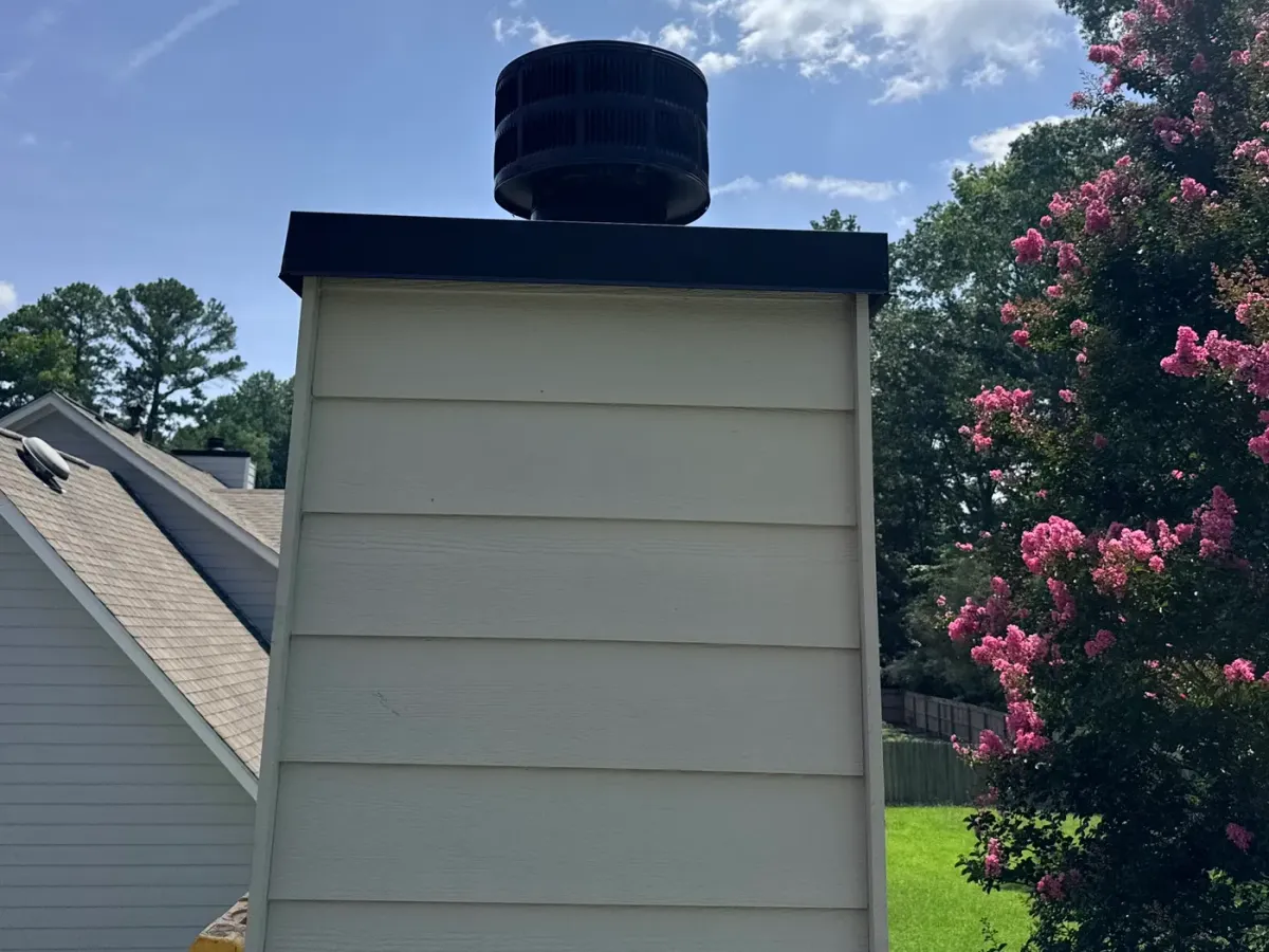 Sided chimney chase with a newly fitted black metal chase cover and round cap on top, photographed from the roof with the adjoining roof slope and a flowering crepe myrtle behind.
