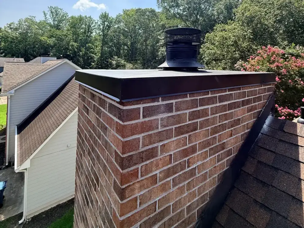Brick chimney with a new dark metal chimney crown and cap fitted on top, fresh counterflashing sealed where the brickwork meets the shingles, and a neighbouring roofline behind.