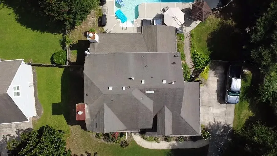 Aerial view of a completed charcoal architectural shingle roof on a large multi-hip home, with even colour across every slope, clean ridge and hip lines, and new vents and pipe boots, a swimming pool and patio beyond.