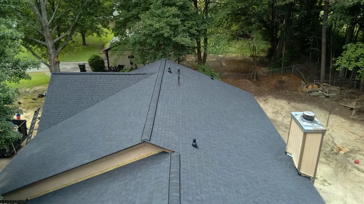 Aerial view of a completed charcoal architectural shingle roof on a large hip-roofed home, showing clean ridge lines, valleys, new pipe boot flashing and a capped chimney.