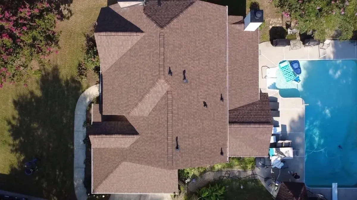 The finished roof on the same home from above: new driftwood-brown architectural shingles, continuous ridge vent along every ridge line, and new black pipe boot flashing at each penetration.