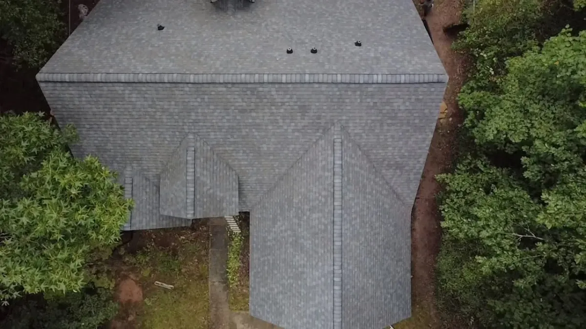 Completed weathered-grey architectural shingle roof on a home surrounded by mature trees, photographed from above, with clean hip and ridge lines and new pipe boot flashing.