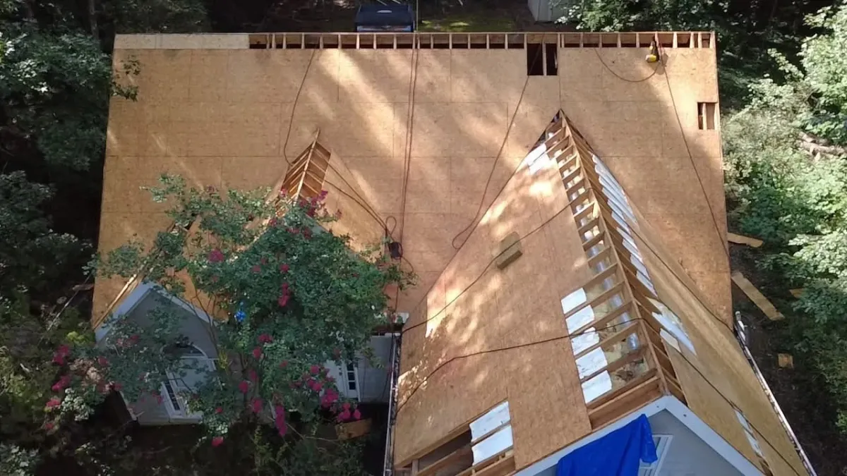 Aerial view of a roof stripped past the shingles to structure, with sections of decking removed entirely and the rafters and insulation open to the sky while fresh sheathing is laid.