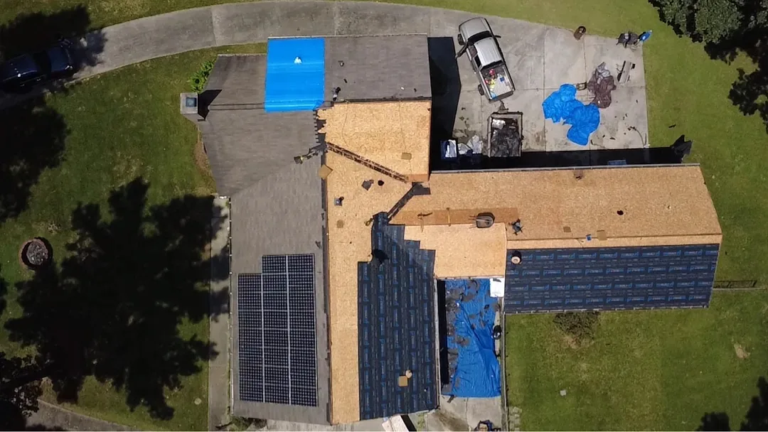 Top-down aerial of a ranch home mid-job: several slopes stripped to bare decking, underlayment going down, tarps on the ground and a dump trailer in the driveway.