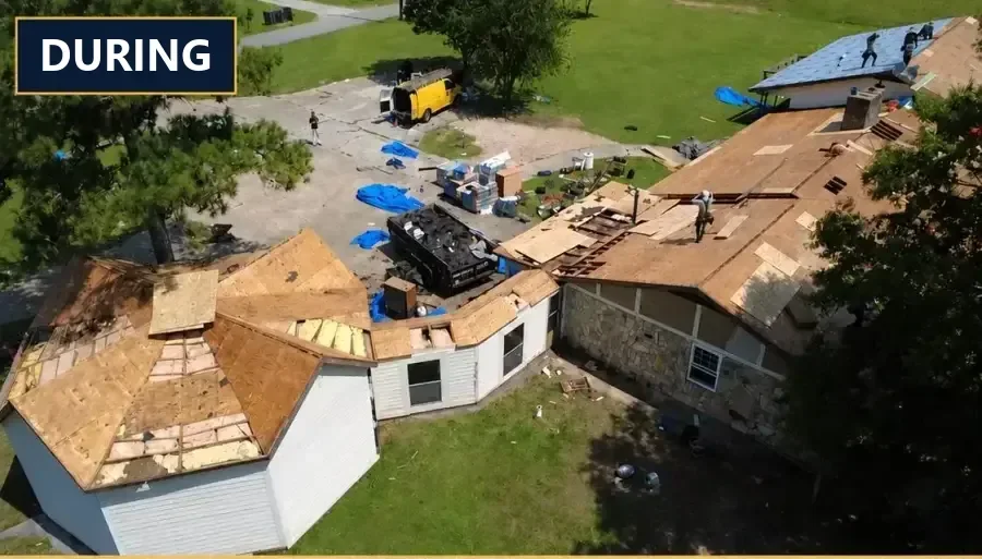 Aerial view of a home mid tear-off: roof decking exposed across every slope, a loaded dump trailer and materials staged in the driveway, and crew working the ridge.