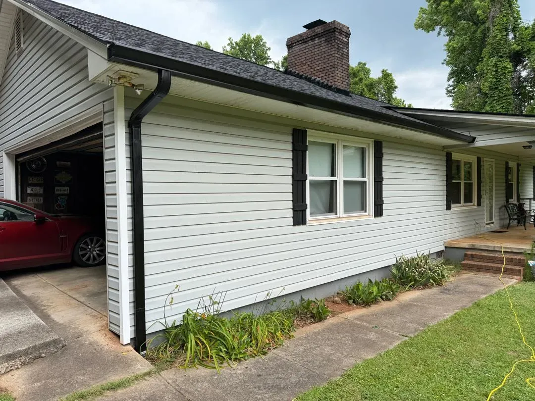 Newly installed black seamless gutters and downspout running along the roofline and corner of a white ranch home, with new shingle roof and drip edge visible above.