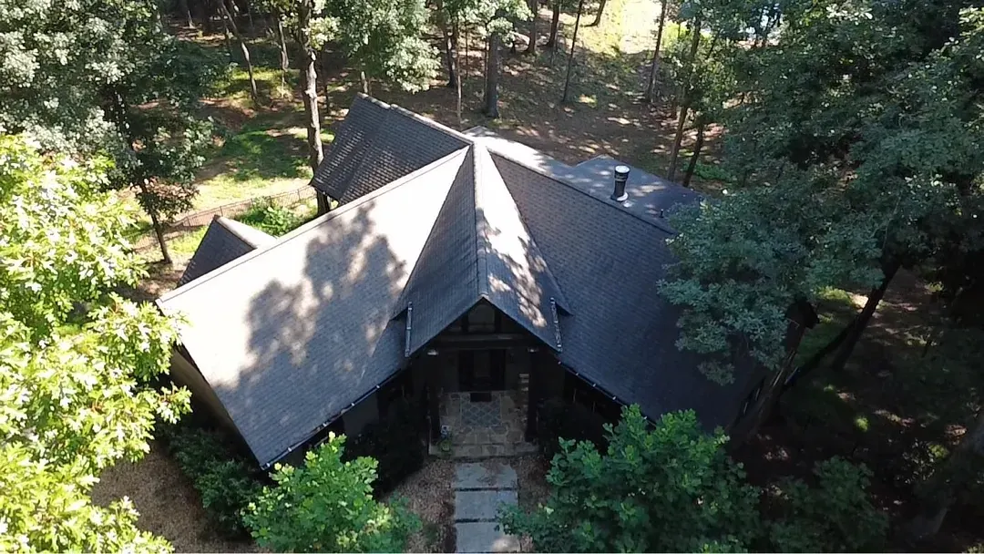 Aerial view of a dark modern home in the woods before the work, still wearing its original dark architectural shingle roof.