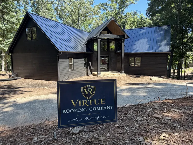 The same home from the driveway: black standing seam roof over dark siding, with a Virtue Roofing Company yard sign in the foreground.