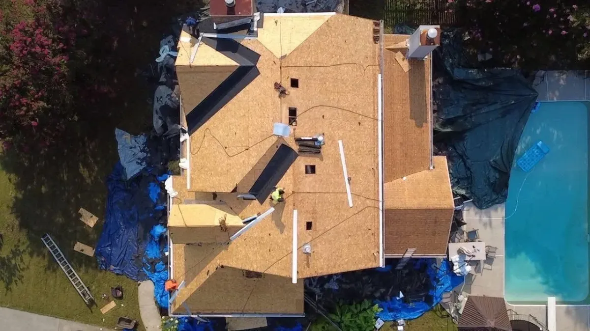 The same home mid tear-off, photographed from above with the roof stripped to bare decking, ice-and-water membrane laid into the valleys, a crew member in a high-visibility vest on the ridge, and tarps covering the pool, patio and planting beds below.